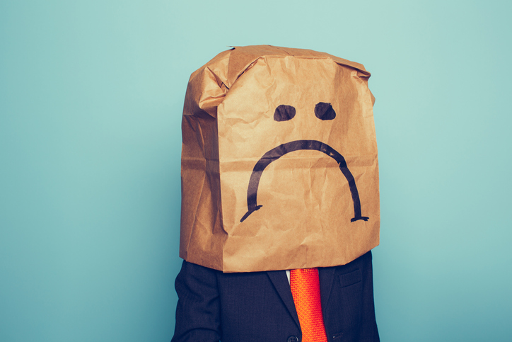 A young boy dressed as a businessman wears a paper bag with a sad face on it The economy has pushed his business down. He sits at his desk with retro computer and phone.